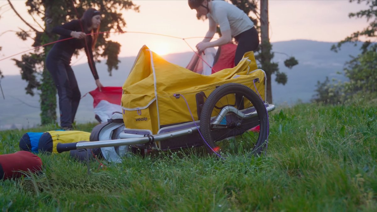 Remorque vélo jaune Veolo en bivouac avec deux cyclistes installant une tente au coucher du soleil.