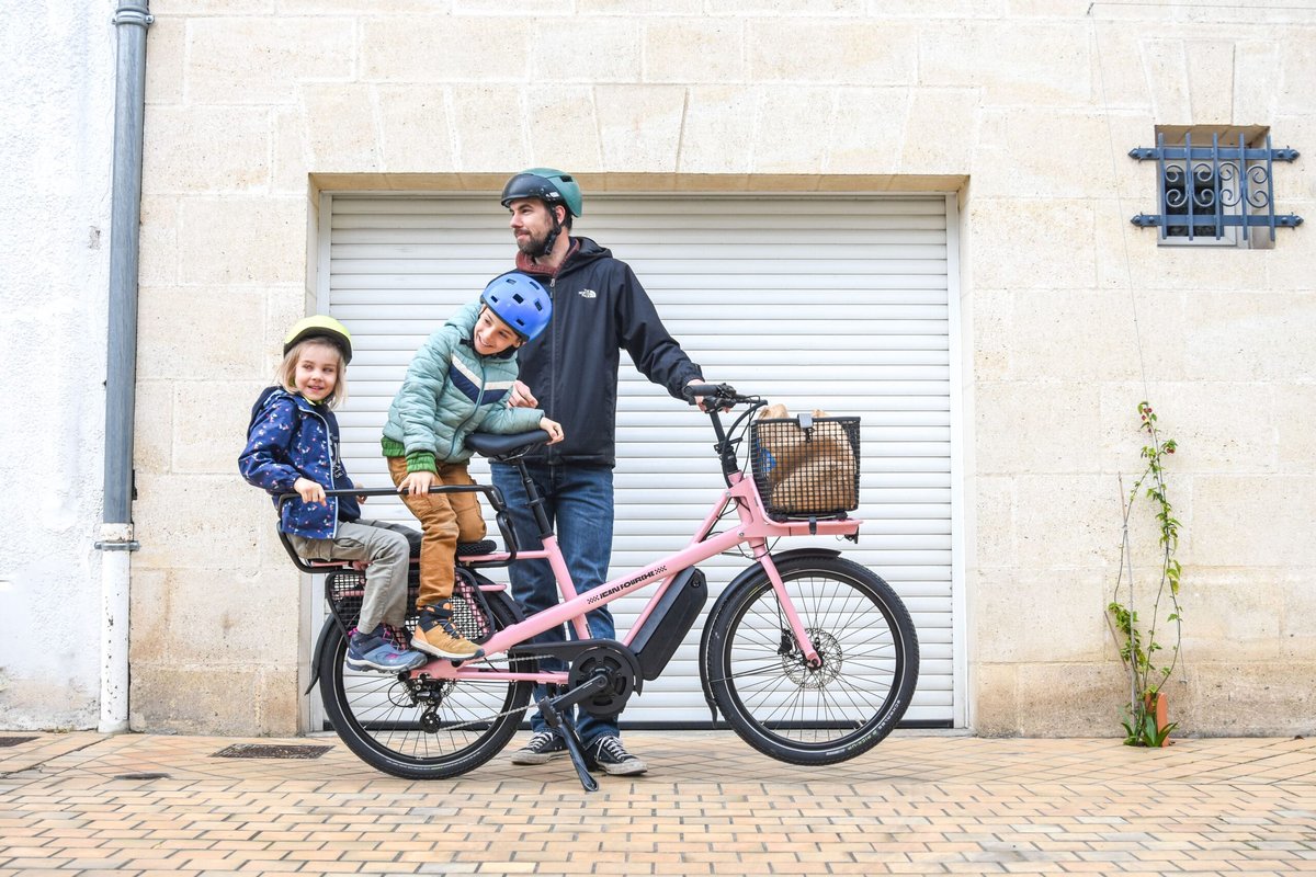 Père et ses deux enfants casqués autour d'un vélo cargo électrique en ville, prêts pour la rentrée