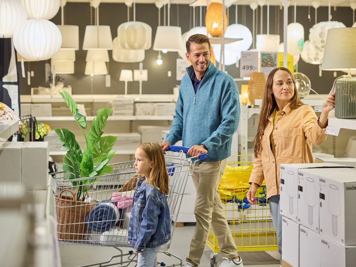 Une famille pousse un chariot rempli d’articles dans le rayon luminaires d’un magasin Ikea, entre suspensions blanches et cartons de lampes.