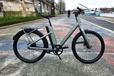 Un vélo électrique Cowboy à l'arrêt sur une rue pavée, avec un cycliste urbain en tenue de vélotaf