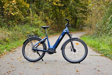 Vélo électrique O2feel Duma bleu posé sur une route forestière en automne