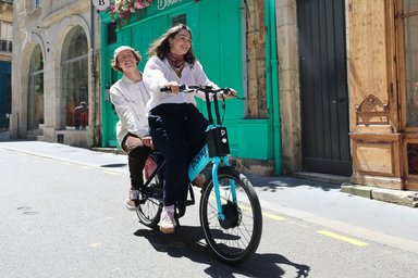 Un couple tout sourire à deux sur un vélo électrique Pony dans une rue en ville.