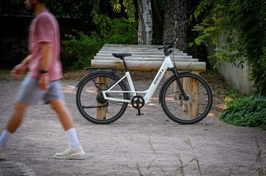 Vélo électrique blanc Ananda avec cadre bas et moteur arrière innovant, stationné devant une structure en bois.