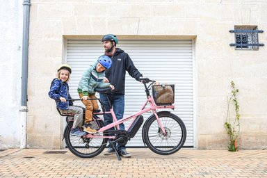 Père et ses deux enfants casqués autour d'un vélo cargo électrique en ville, prêts pour la rentrée