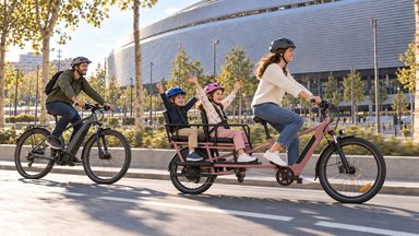 Femme sur un vélo cargo longtail rose avec deux enfants casqués a l'arriere, un homme suit en velo electrique