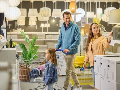 Une famille pousse un chariot rempli d’articles dans le rayon luminaires d’un magasin Ikea, entre suspensions blanches et cartons de lampes.