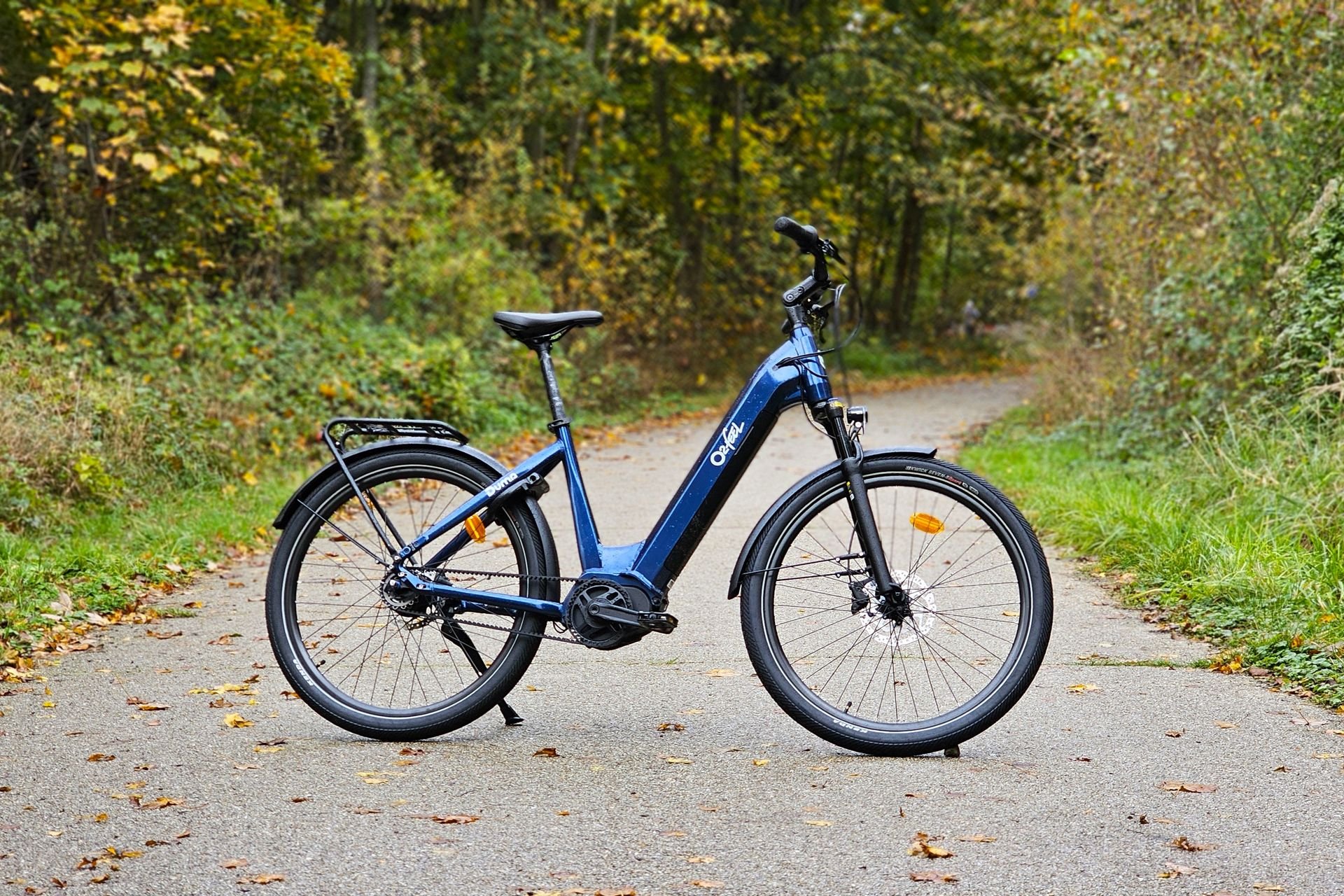 Vélo électrique O2feel Duma bleu posé sur une route forestière en automne