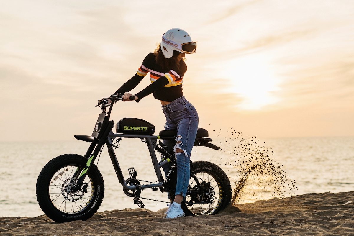 Une femme pilote un vélo électrique Super73 RX puissant sur une plage de sable au coucher du soleil.